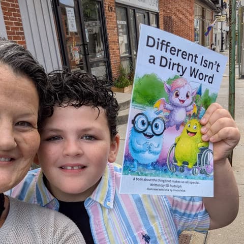Eli and his mom, the book's co-author, cheek to cheek and smiling on a small-town sidewalk while Eli holds up a copy of Different Isn't a Dirty Word.