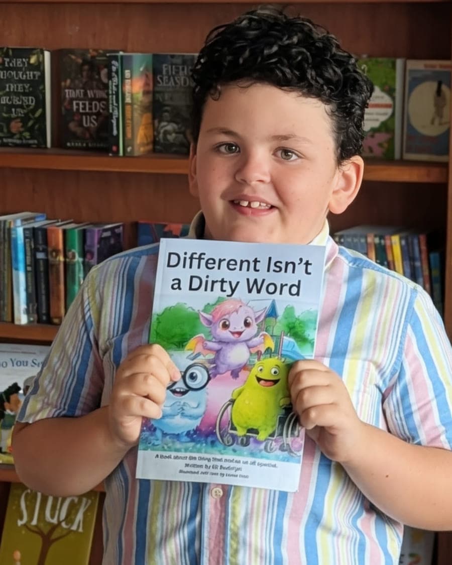 Eli, a young boy with short curly dark hair, wearing a pastel striped button-down shirt and smiling in front of a bookshop's shelves while holding up a copy of his book Different Isn't a Dirty Word.